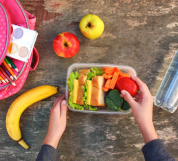 Healthy and unhealthy school snacks and snacks for kids: Sandwiches, fruits and vegetables in food box, backpack and bottled water on table.