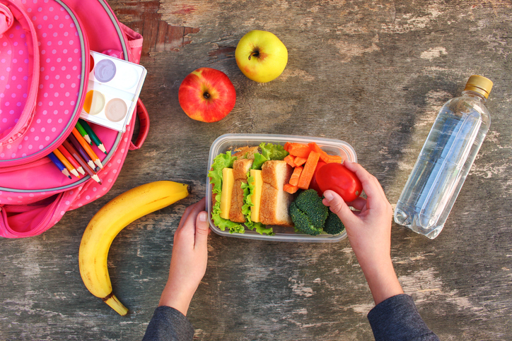 Healthy and unhealthy school snacks and snacks for kids: Sandwiches, fruits and vegetables in food box, backpack and bottled water on table.